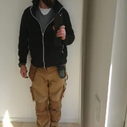 Mężczyzna w odzieży roboczej, okularach przeciwsłonecznych i czapce z daszkiem, trzymający telefon na tle białej ściany i jasnej drewnianej podłogi.