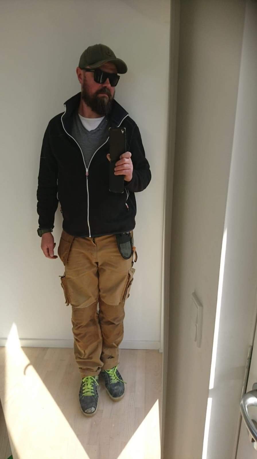 Mężczyzna w odzieży roboczej, okularach przeciwsłonecznych i czapce z daszkiem, trzymający telefon na tle białej ściany i jasnej drewnianej podłogi.