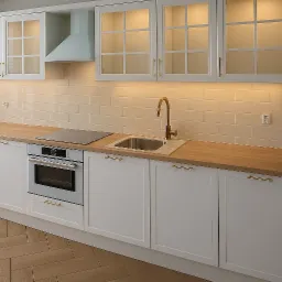 Jasna kuchnia z białymi szafkami, drewnianym blatem i złotymi uchwytami. Uwagę zwraca minimalistyczny okap i płytki w odcieniach beżu, podkreślone ciepłym oświetleniem.