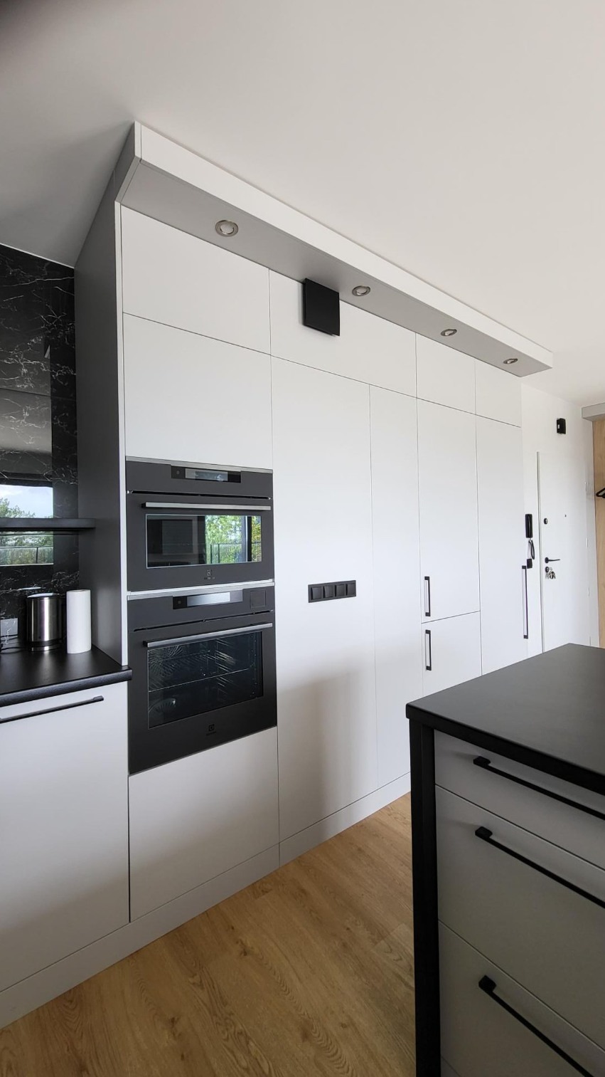 Biała zabudowa kuchenna z piekarnikiem i mikrofalówką Electrolux, czarne uchwyty, marmurowy backsplash, drewniana podłoga, nowoczesny design.
