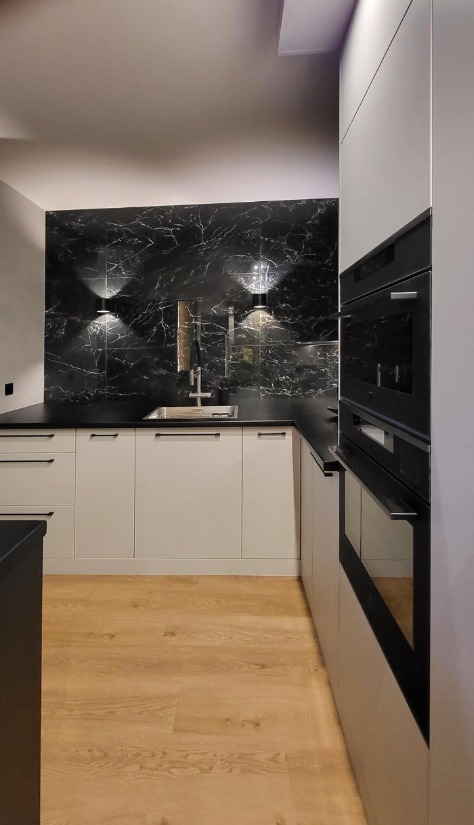 Nowoczesna kuchnia z marmurowym panelem, oświetleniem punktowym i zabudową piekarnika. Jasne fronty szafek i drewniana podłoga tworzą elegancki kontrast.