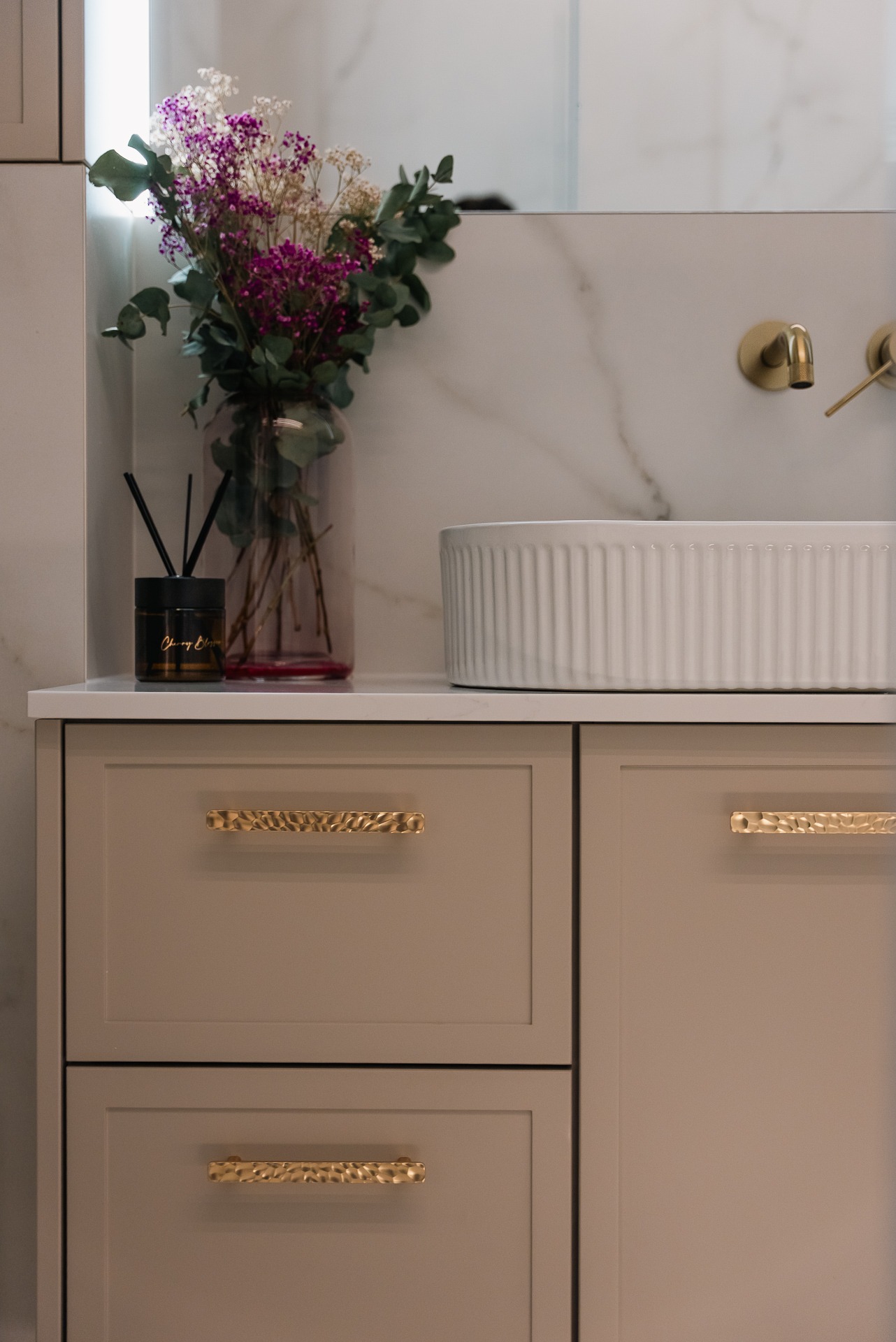 Beżowa szafka łazienkowa z frezowanymi frontami i złotymi uchwytami. Na blacie wazon z kwiatami i dyfuzor zapachowy. Biała umywalka nablatowa.