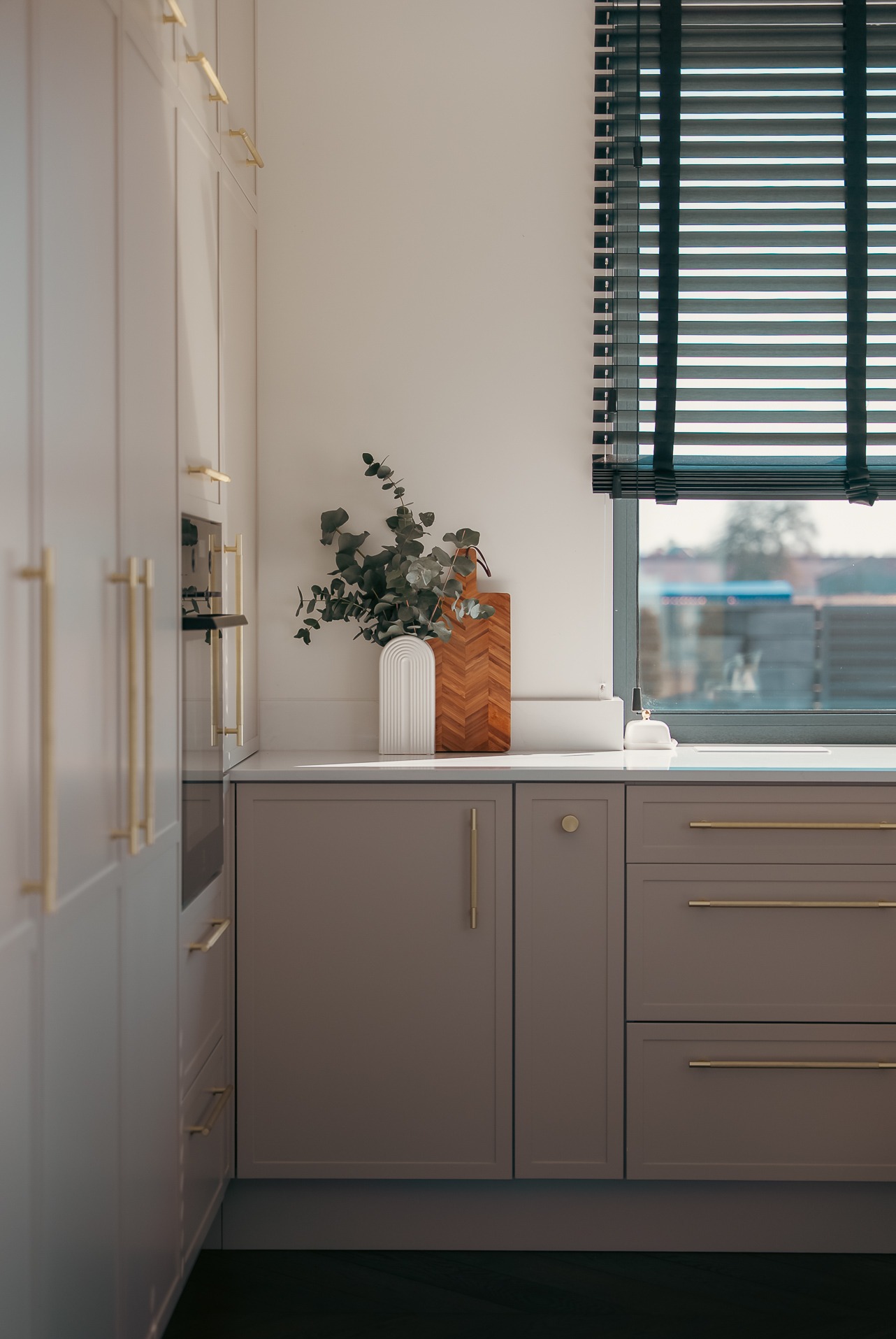 Szare szafki kuchenne z złotymi uchwytami, biały blat, wazon z eukaliptusem i deska do krojenia. Okno z żaluzjami w tle. Minimalistyczny design.