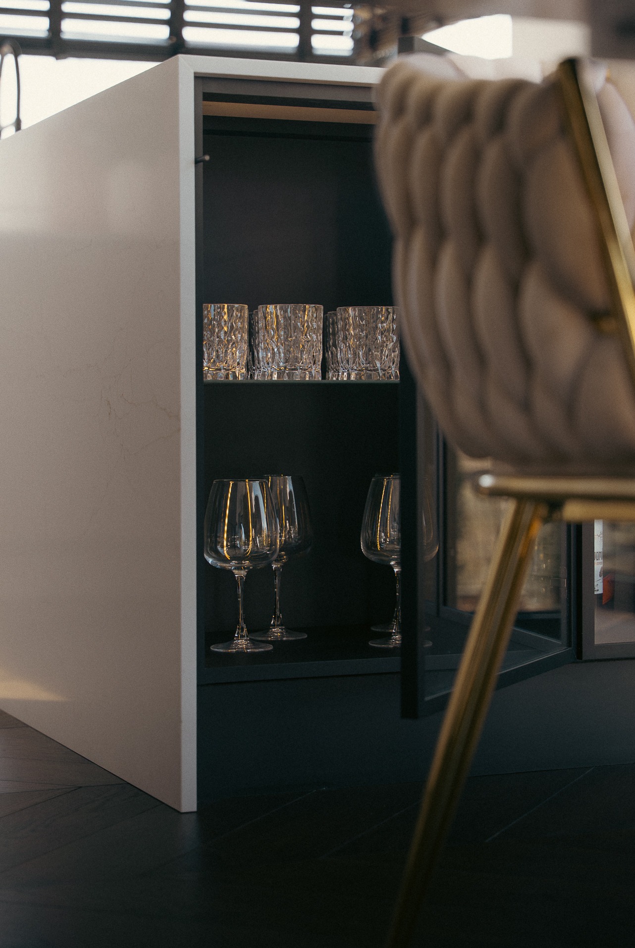 Minimalistyczna szafka kuchenna z otwartą półką, na której stoją szklanki i kieliszki. W tle fragment krzesła z tapicerowanym oparciem.