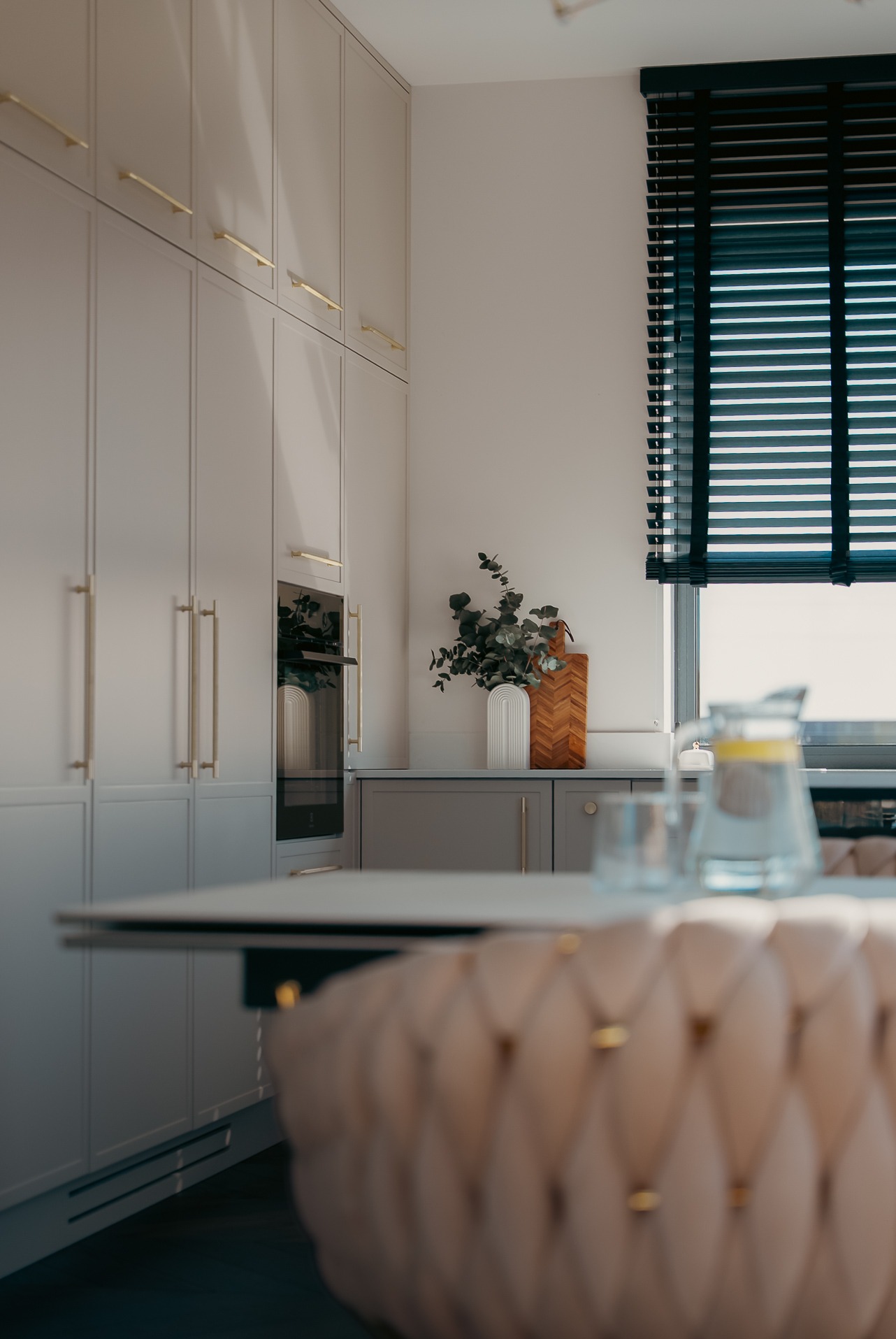 Elegancka kuchnia w odcieniach szarości z złotymi uchwytami, minimalistycznym wystrojem i drewnianymi akcentami, widziana z perspektywy stołu z tapicerowanym krzesłem.