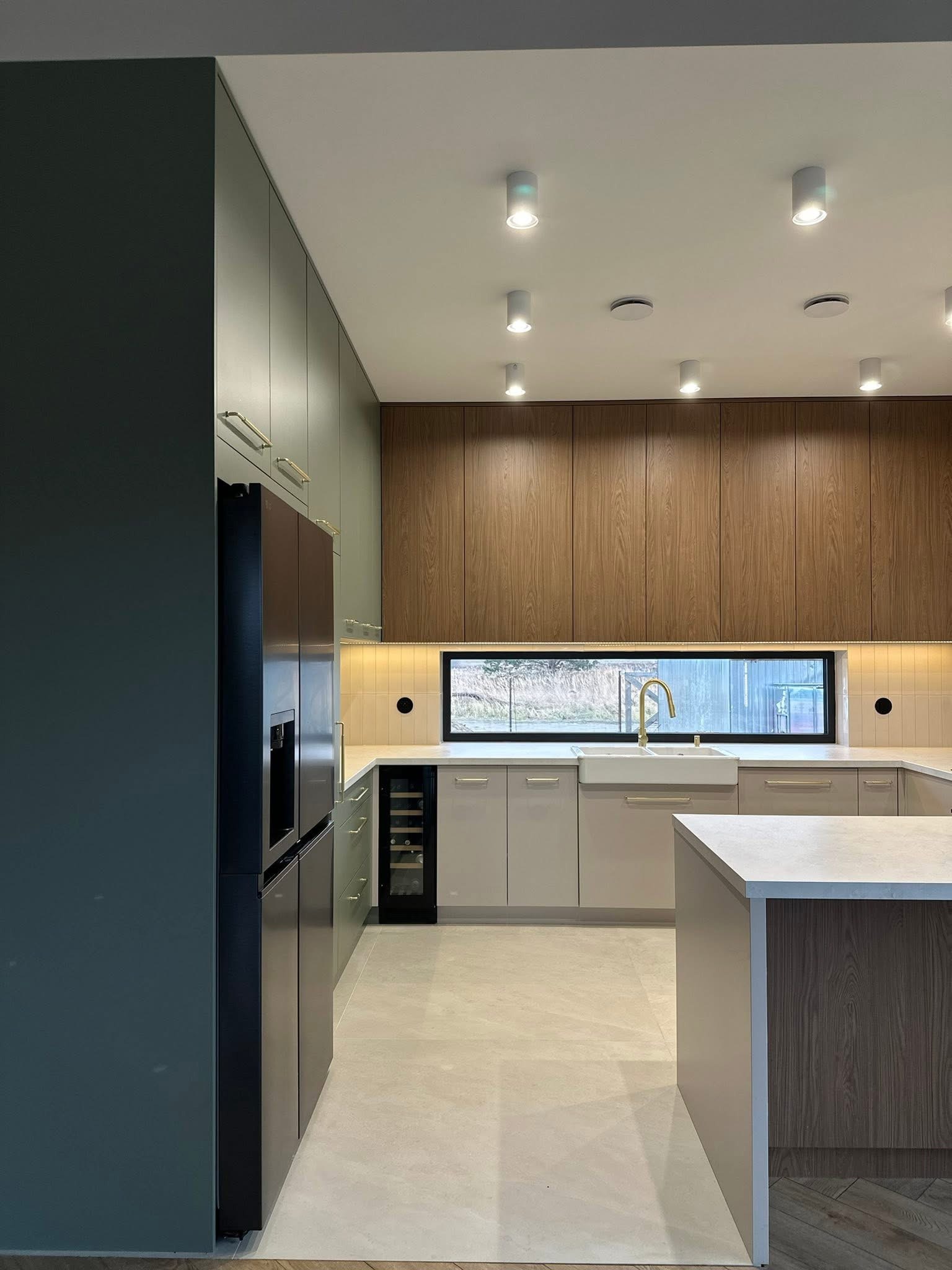 Nowoczesna kuchnia z wyspą, fronty w odcieniach szarości i drewna, złota armatura, oświetlenie punktowe. Widoczna lodówka z kostkarką i winiarka.