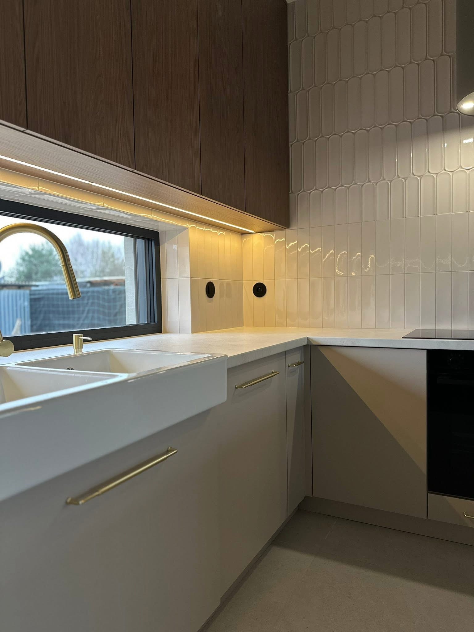 Nowoczesna kuchnia z białym, głębokim zlewozmywakiem pod oknem, złotą armaturą i podświetleniem LED. Uwagę zwracają beżowe fronty szafek i płytki w kształcie migdałów.