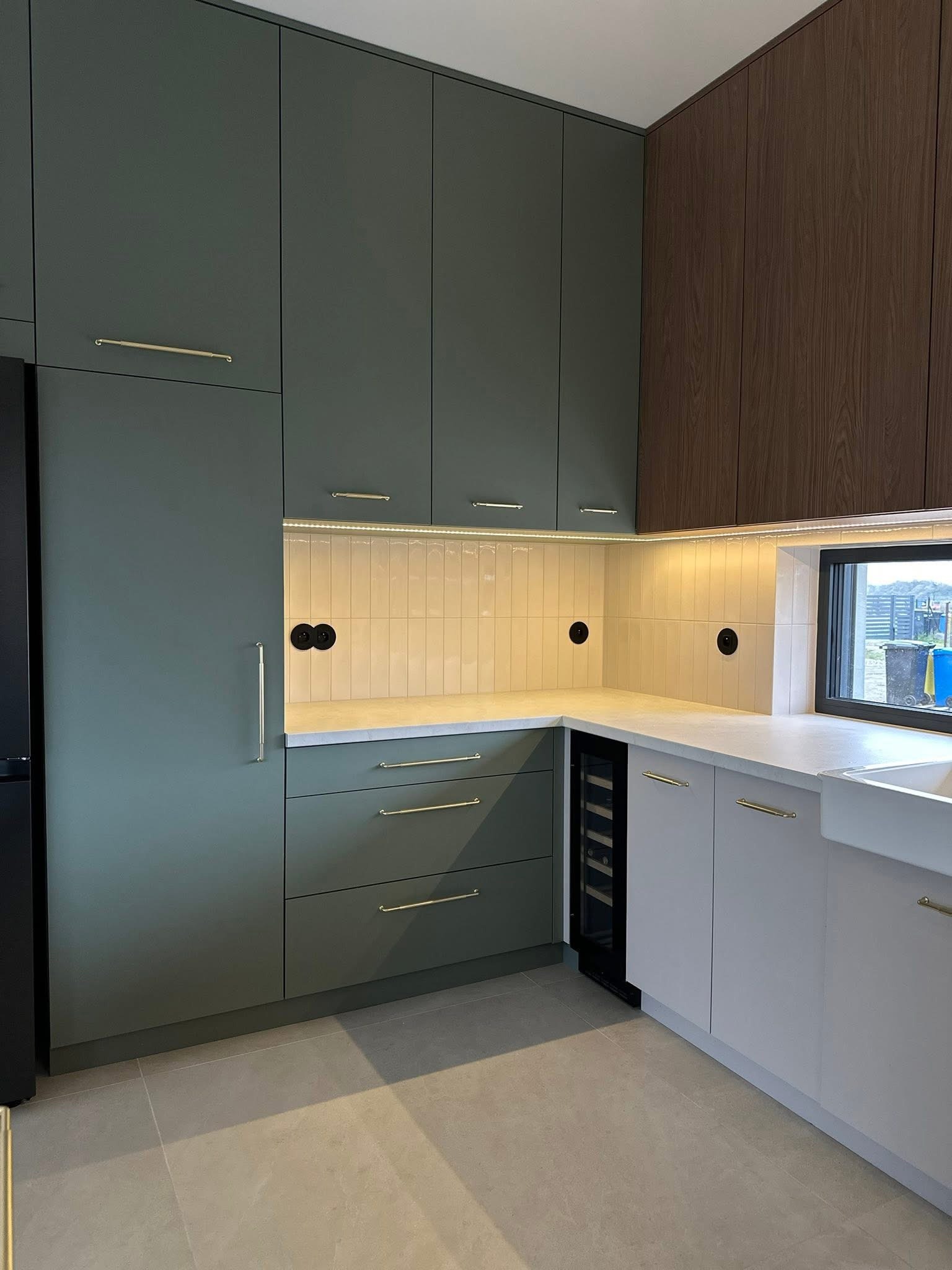 Nowoczesna kuchnia z zielonymi i drewnianymi szafkami, białym blatem, oświetleniem LED i czarnymi gniazdkami. Widoczna zabudowa z lodówką i szafką na wino.