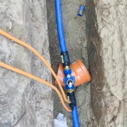 Wykop z widocznymi niebieskimi rurami wodociągowymi połączonymi z pomarańczową studzienką rewizyjną, pomarańczowe przewody w tle, biała szmatka na ziemi.