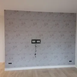 Ściana z tapetą imitującą cegłę, przygotowana do montażu telewizora, widoczne gniazdka elektryczne i uchwyt na ścianie z wystającymi przewodami.