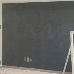 Ściana pokryta ciemną, strukturalną gładzią z geometrycznym wzorem, widoczna drabina malarska, niezabezpieczone gniazdka elektryczne i listwa przypodłogowa.