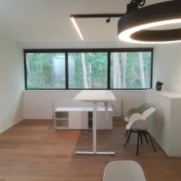 Jasne, minimalistyczne wnętrze biurowe z białymi ścianami, drewnianą podłogą, dużym oknem z widokiem na las, nowoczesnym oświetleniem i białymi meblami.