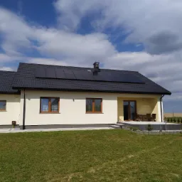 Jednorodzinny dom z ciemną dachówką i panelami fotowoltaicznymi na dachu, widok z trawnika w słoneczny dzień z chmurami.