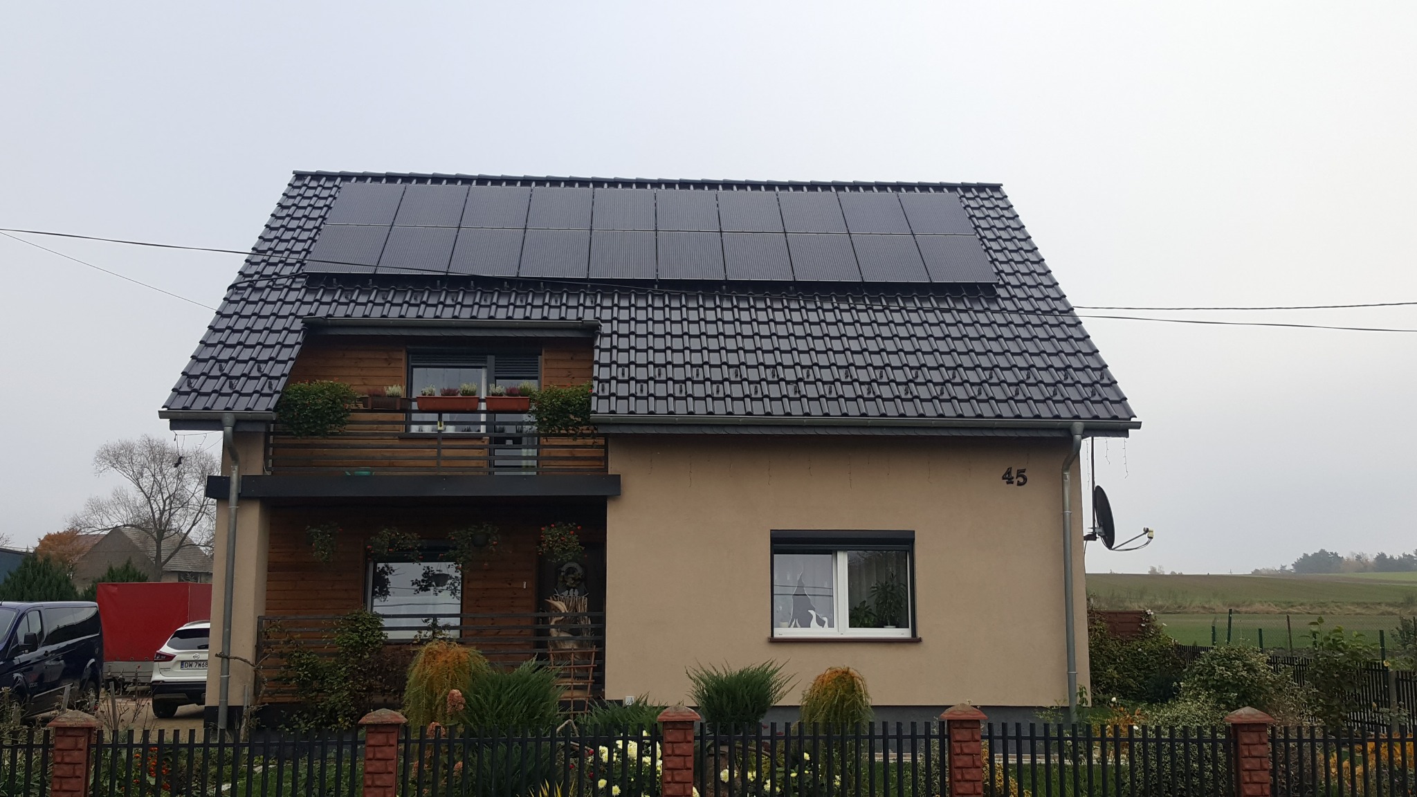 Dom jednorodzinny z ciemną dachówką i panelami fotowoltaicznymi na dachu, widoczny balkon z kwiatami i numer domu '45' w pochmurny dzień.