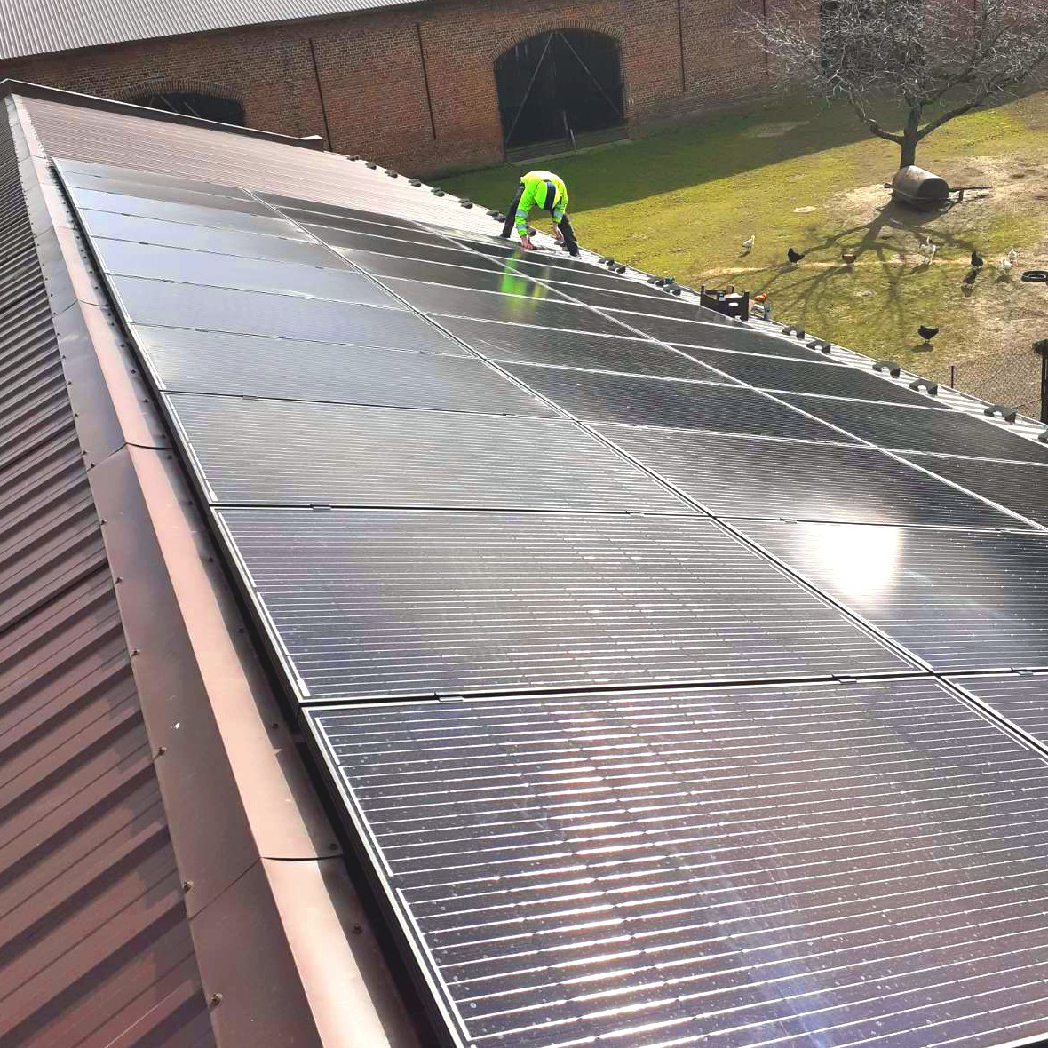 Montaż paneli słonecznych na dachu z blachy trapezowej, pracownik w odblaskowej kamizelce na tle budynku gospodarczego i zielonego trawnika.