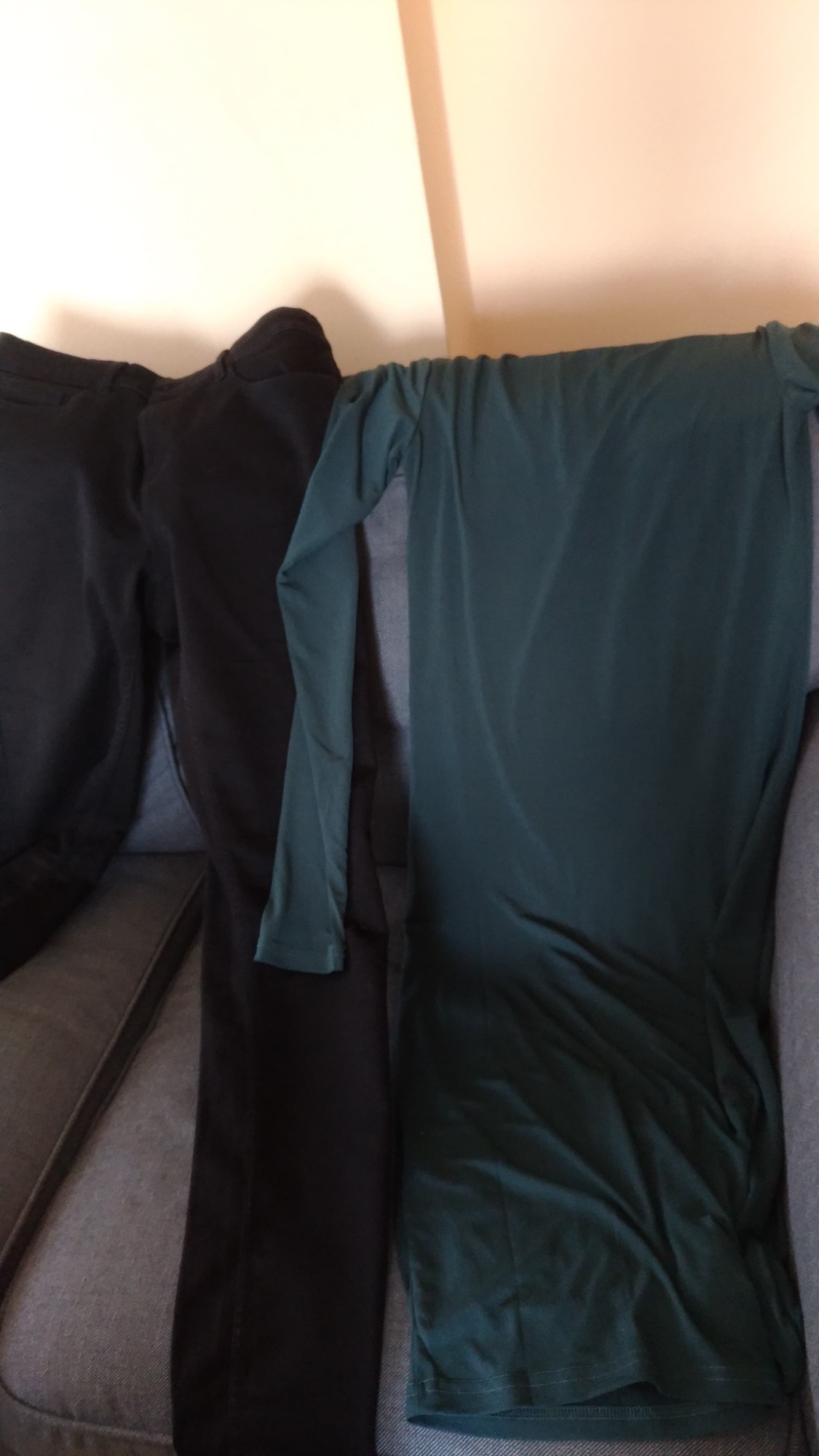 Dwie pary czarnych spodni i ciemnozielona sukienka z długim rękawem, ułożone na szarej kanapie.