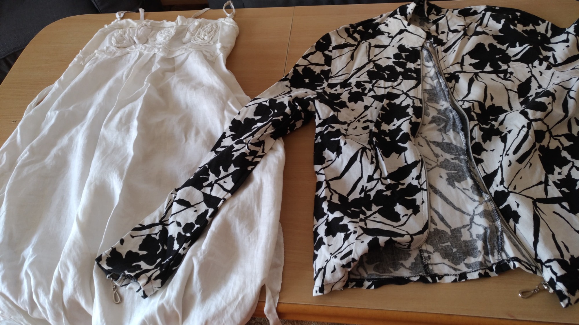 Biała sukienka z aplikacjami kwiatowymi leżąca obok rozpiętej kurtki z wzorem czarno-białych liści na drewnianym stole.