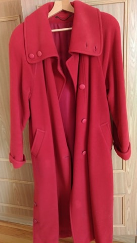 Długi, czerwony płaszcz wełniany z kołnierzem i dużymi guzikami, wiszący na drewnianym wieszaku na tle jasnych drzwi.