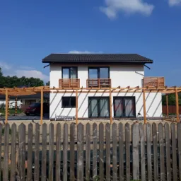 Nowoczesny dom z białymi ścianami i czarnym dachem, z drewnianą konstrukcją pergoli w stanie surowym, widoczną zza drewnianego płotu, na tle błękitnego nieba.