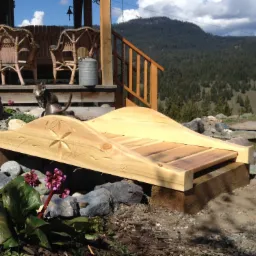 Mały, ozdobny mostek z jasnego drewna, z wyrytym wzorem gwiazdy i kwiatów, prowadzący nad małym oczkiem wodnym; w tle weranda z wiklinowymi fotelami i kotem.