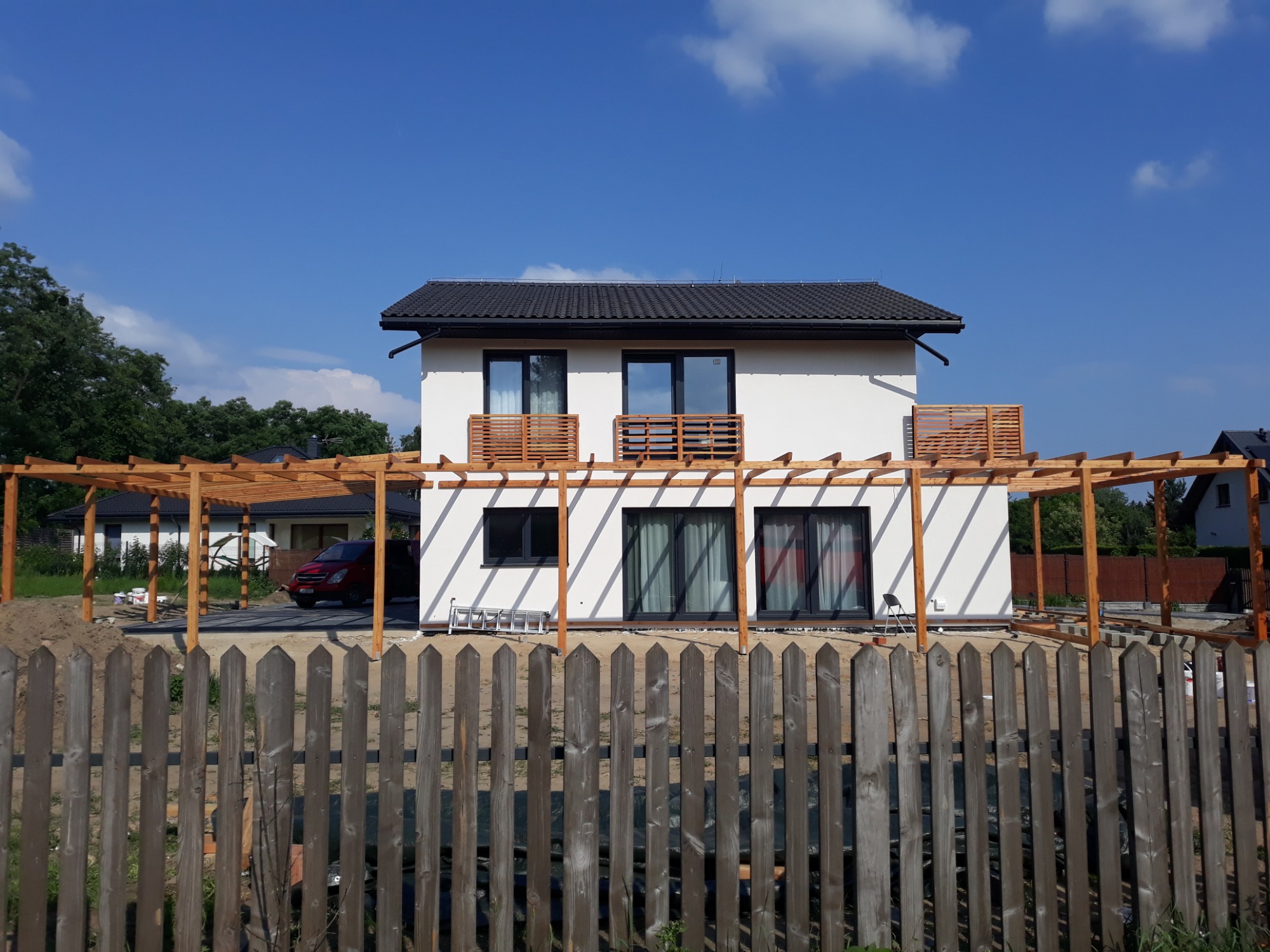 Nowoczesny dom z białymi ścianami i czarnym dachem, z drewnianą konstrukcją pergoli w stanie surowym, widoczną zza drewnianego płotu, na tle błękitnego nieba.