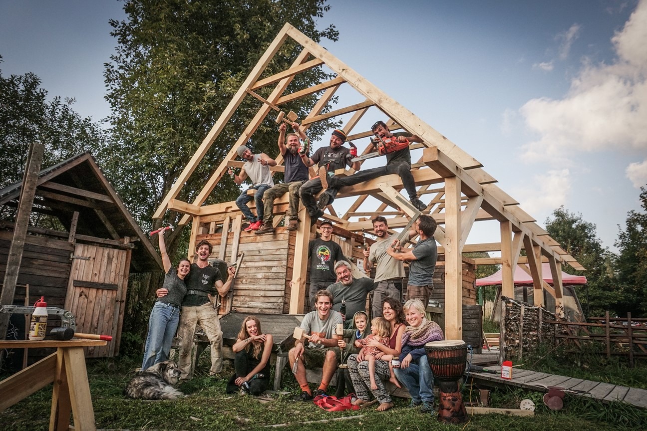 Grupa osób pozuje na tle drewnianej konstrukcji szkieletowej budynku w trakcie budowy, z widocznymi narzędziami i elementami drewnianymi, na zewnątrz w naturalnym otoczeniu z drzewami i zielenią.
