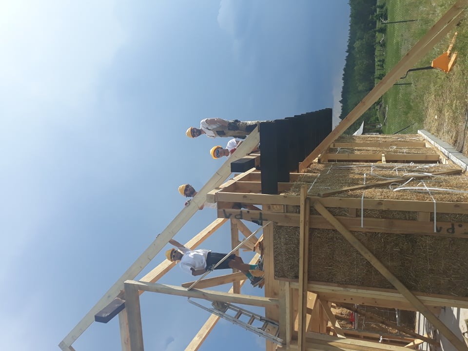 Budowa domu ze słomy: grupa robotników w kaskach montuje drewnianą więźbę dachową na ścianach z kostek słomy, widoczne narzędzia i drabina.