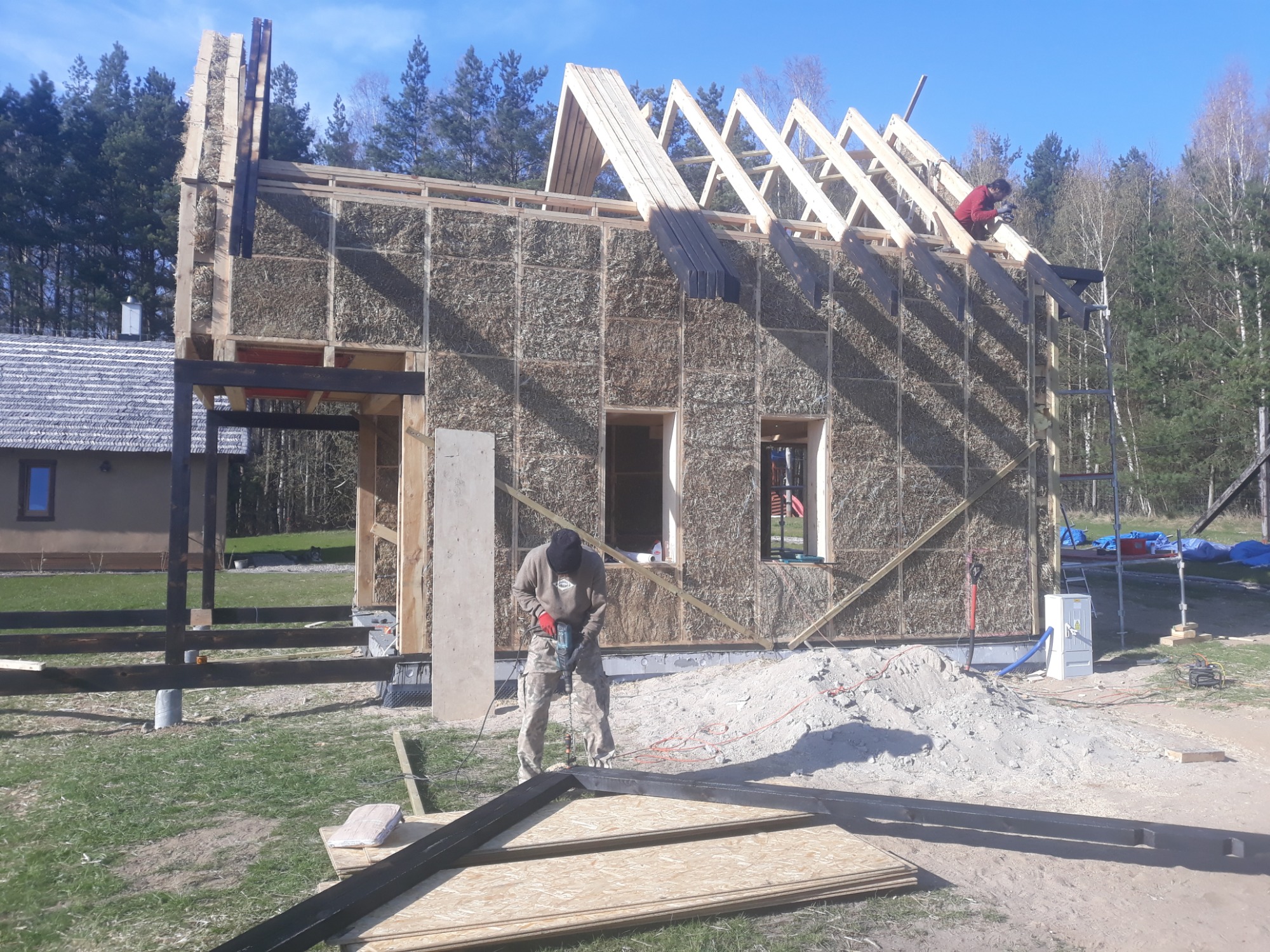 Budowa domu ze ścianami z kostek słomy, widoczna drewniana konstrukcja dachu i pracownicy podczas montażu więźby dachowej.