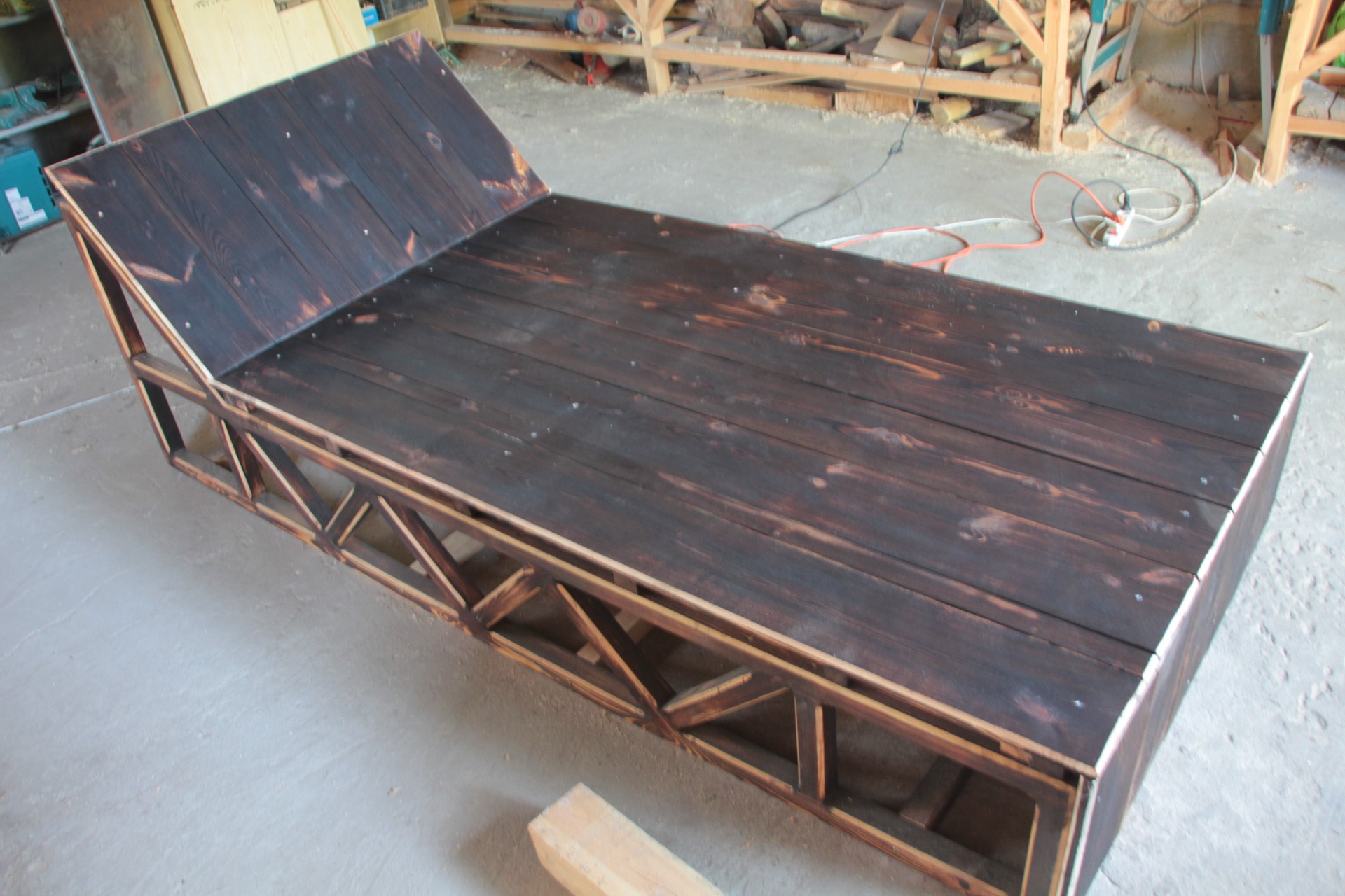 Konstrukcja leżanki z ciemnego, bejcowanego drewna z widoczną ramą nośną z jasnego drewna, znajdująca się w warsztacie.