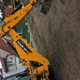 Żółta koparka JCB z łyżką na tle świeżo wykopanej ziemi, z widocznymi domami w tle.