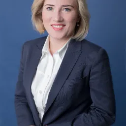 Portret uśmiechniętej blondynki w granatowej marynarce i białej koszuli na jednolitym niebieskim tle.