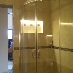 Kabina prysznicowa z przezroczystego szkła z chromowanymi uchwytami, zamontowana w łazience wykończonej beżowymi płytkami z dekoracyjnym pasem mozaiki. W tle widoczne niedokończone prace remontowe.