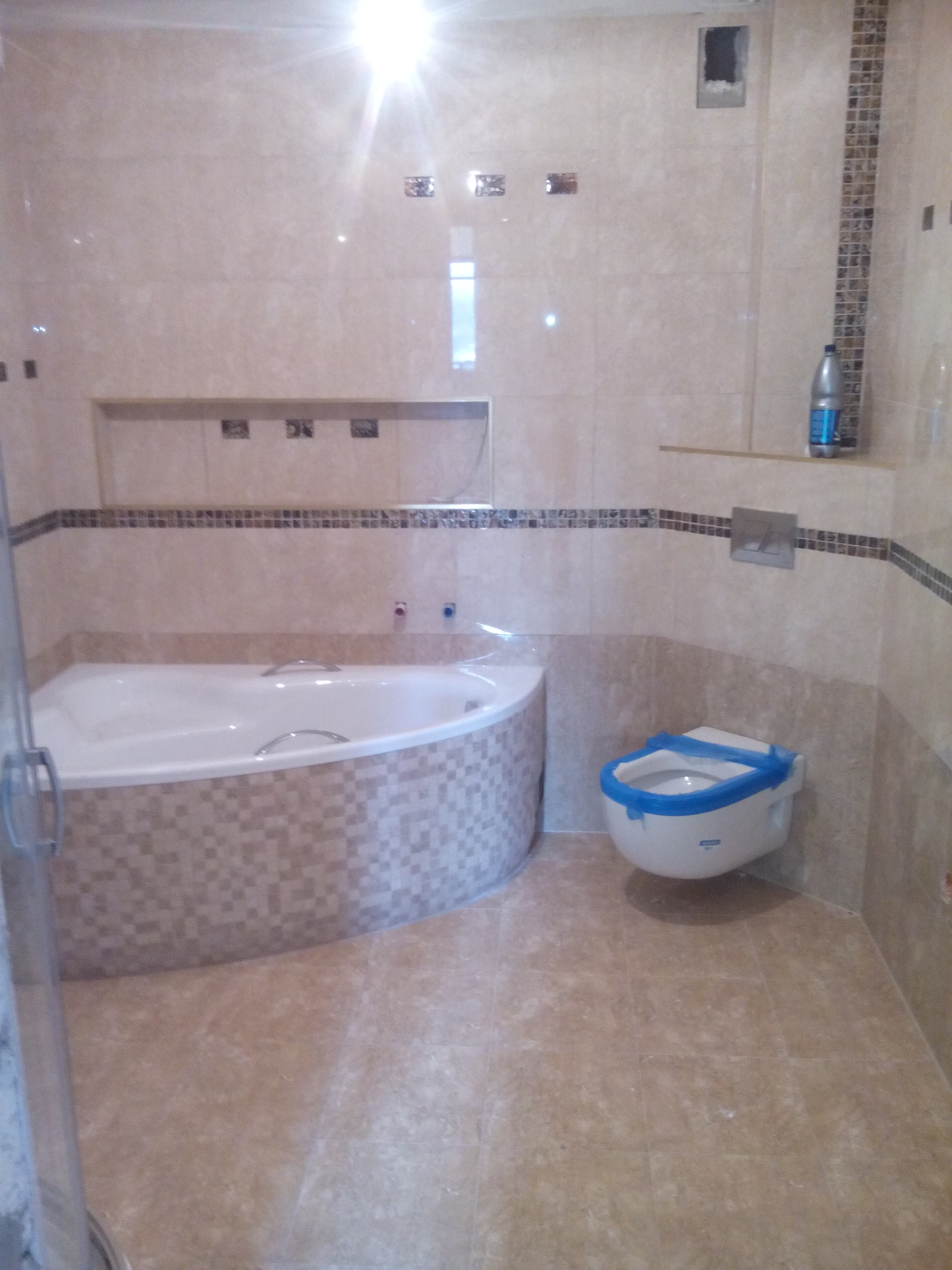 Wykończona łazienka z beżowymi płytkami, narożną wanną obudowaną mozaiką i podwieszanym sedesem zabezpieczonym niebieską folią ochronną.