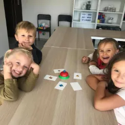 Czwórka uśmiechniętych dzieci siedzi przy stole, grając w grę edukacyjną z kartami i dzwonkiem, w tle półka z książkami i dekoracjami w klasie.