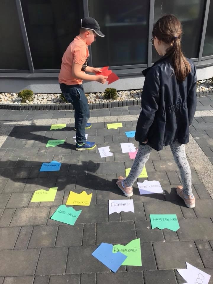 Dwoje dzieci bawi się na zewnątrz, układając kolorowe kartki z nazwami zawodów i miejsc na chodniku, tworząc interaktywną grę edukacyjną.