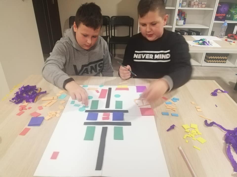 Dwóch chłopców układa geometryczną kompozycję z kolorowych elementów na dużej kartce papieru, otoczeni resztkami materiałów na jasnym stole.