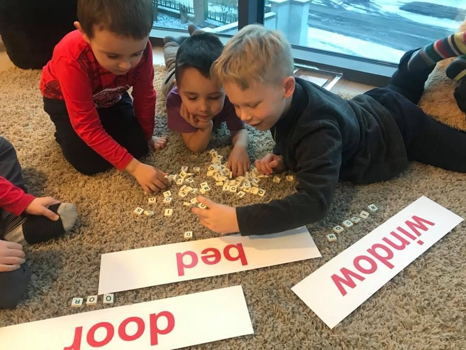 Grupa dzieci na dywanie układa słowa z literkowych kostek, obok leżą kartki z napisanymi słowami: door, bed, window.