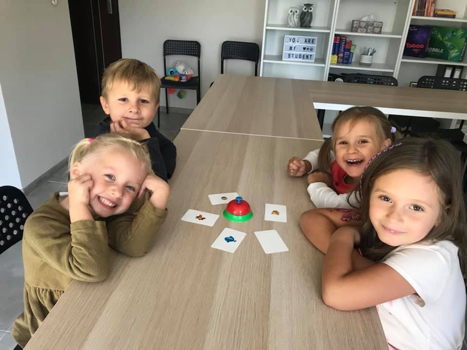 Czwórka uśmiechniętych dzieci siedzi przy stole, grając w grę edukacyjną z kartami i dzwonkiem, w tle półka z książkami i dekoracjami w klasie.