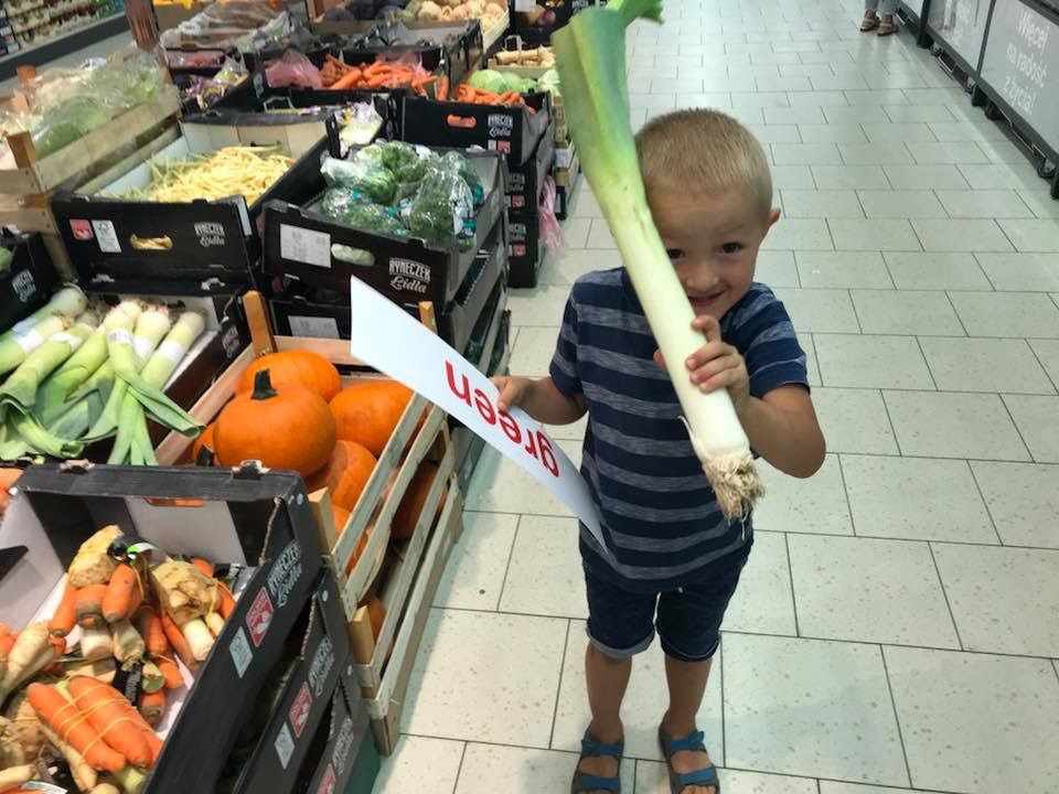 Uśmiechnięty chłopiec w sklepie spożywczym trzyma w rękach duży por i kartkę z napisem 'Green', stojąc obok skrzynek z warzywami.