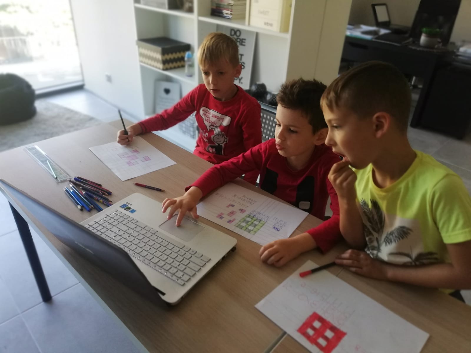 Trzej chłopcy w różnym wieku siedzą przy stole, rysują na papierze i korzystają z laptopa, prawdopodobnie ucząc się lub bawiąc.