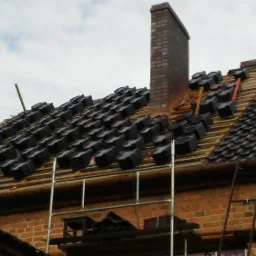 Częściowo pokryty dach z czarną dach&oacute;wką, widoczny komin z cegły, rusztowanie i wiązki dach&oacute;wek czekające na ułożenie.