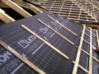 Układanie membrany dachowej z nadrukiem siatki i logo producenta na krokwiach, widoczne łaty dachowe w trakcie montażu.