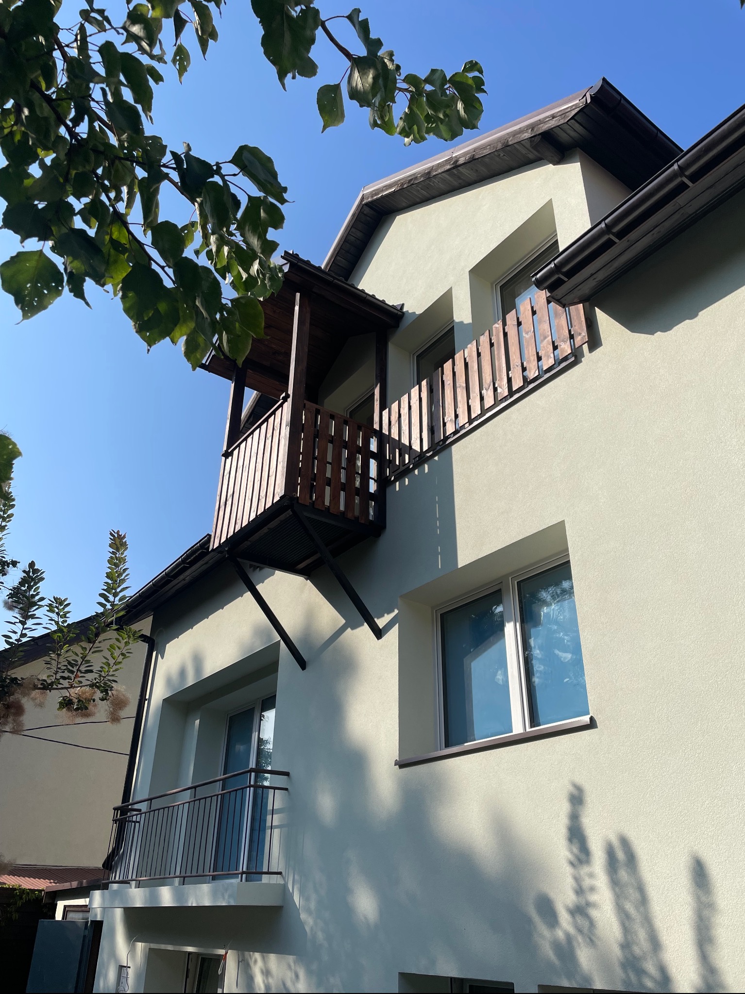 Jasnozielony budynek z dwoma balkonami, jeden z drewnianą balustradą i czarną konstrukcją wsporczą, drugi z metalową balustradą, sfotografowany w słoneczny dzień z fragmentem drzewa w lewym górnym...