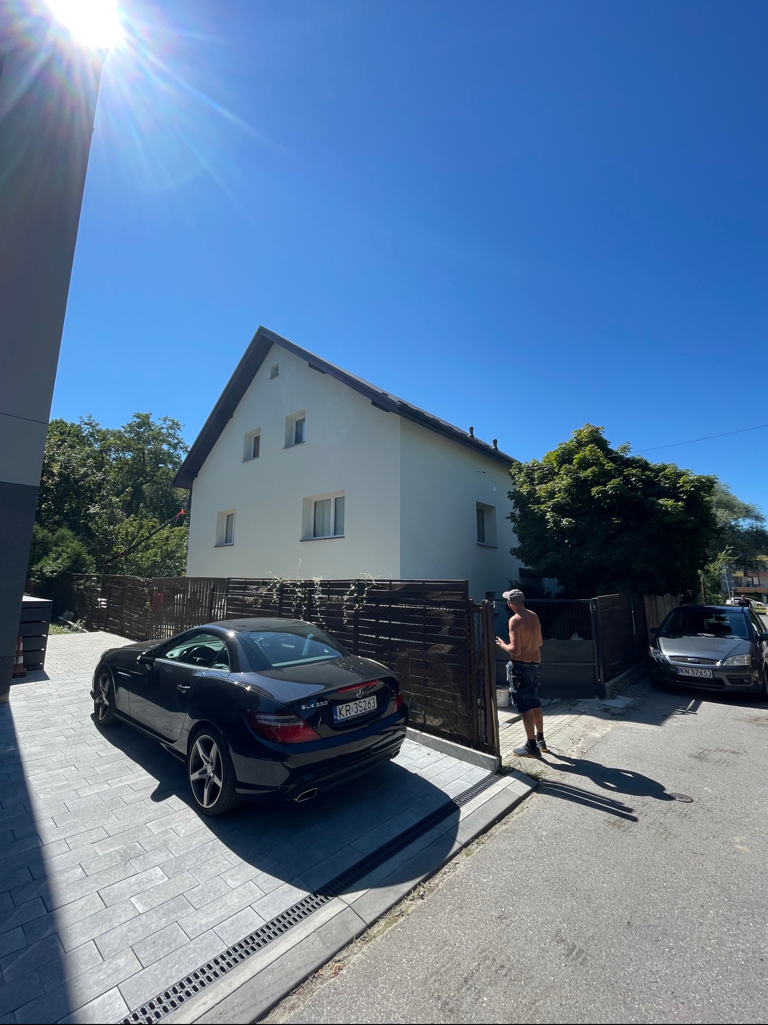 Biały dom jednorodzinny z ciemnym dachem, widziany z perspektywy ulicy, z zaparkowanym czarnym Mercedesem SLK 350 i szarym Fordem Focus. Mężczyzna bez koszulki stoi przy bramie wjazdowej.