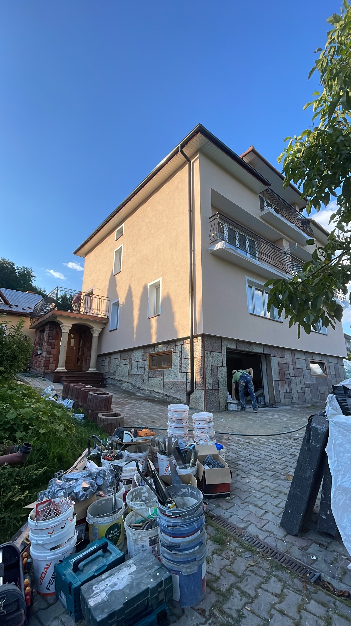 Dwupiętrowy dom z beżową elewacją i kamienną podmurówką, widoczne balkony z ozdobnymi balustradami, w tle błękitne niebo, na pierwszym planie bałagan z wiaderkami po farbach i narzędziami...
