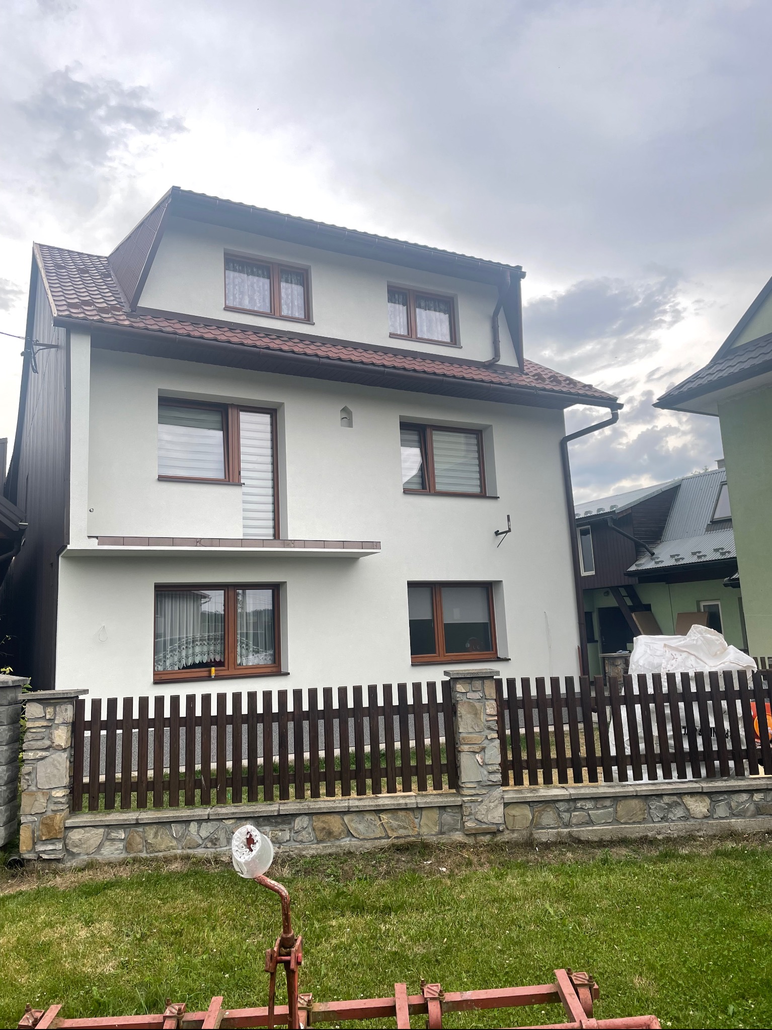 Dwukondygnacyjny dom z jasną elewacją, brązowymi oknami i dachem z czerwonej dachówki, widziany zza drewnianego płotu na kamiennej podmurówce; na pierwszym planie fragment czerwonego sprzętu...