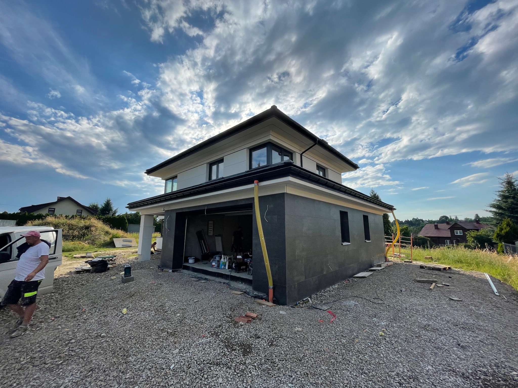 Dwukondygnacyjny dom w trakcie prac elewacyjnych, z widocznym oknem garażowym, szarą elewacją, podsypką żwirową i pracownikiem obok samochodu dostawczego. W tle błękitne niebo z obłokami.