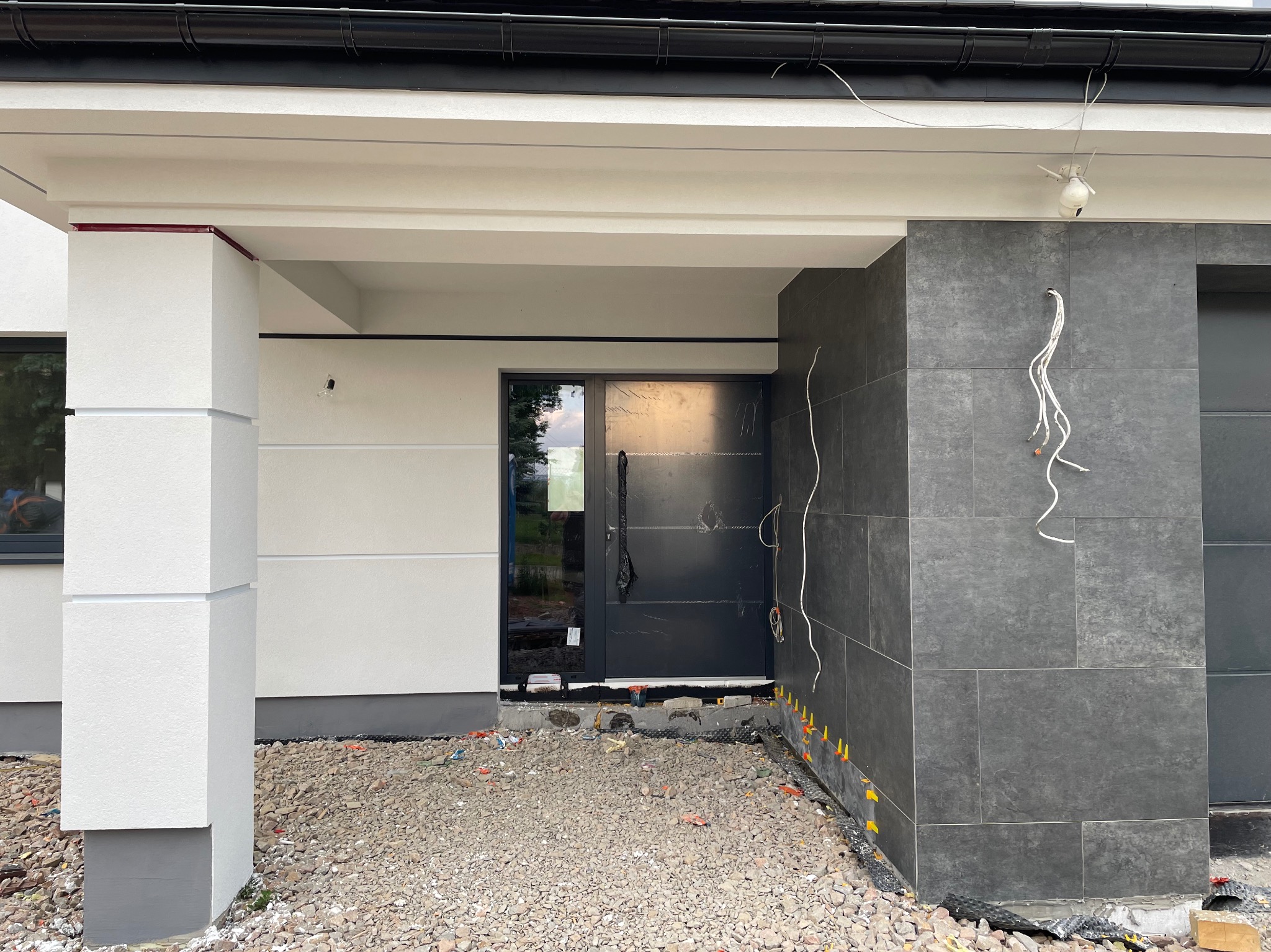 Nowoczesny dom w trakcie wykańczania elewacji, widoczne fragmenty otynkowanej ściany, kolumna z dekoracyjnymi wgłębieniami, ciemne płytki elewacyjne i przygotowane podłoże z kamyków przed wejściem.