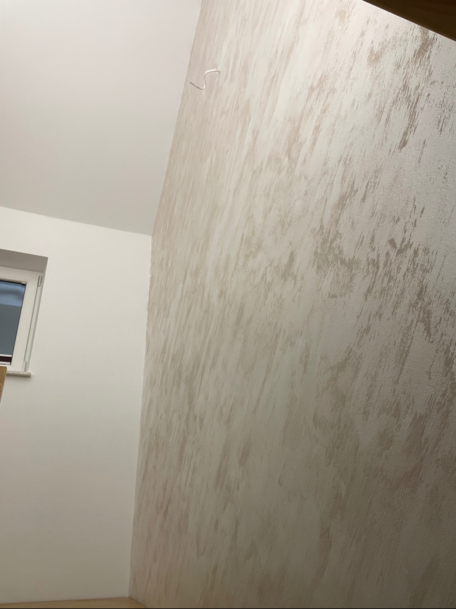 Ściana wewnętrzna z dekoracyjną strukturą w odcieniach beżu i szarości, widoczna z bliska pod kątem, sąsiadująca z gładką, białą ścianą z oknem.