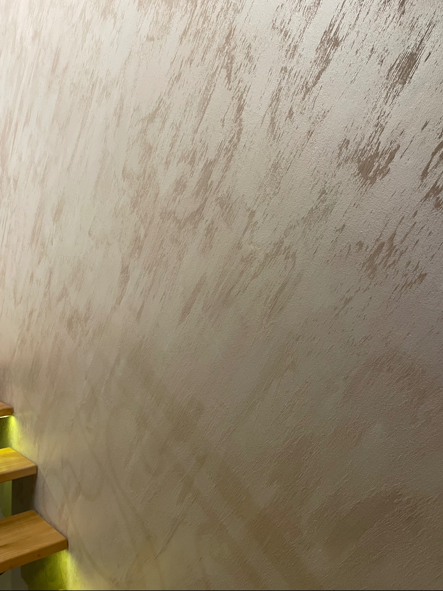 Beżowa ściana z dekoracyjną fakturą przypominającą przecierki, oświetlona od dołu przez ledowe światło schodów z drewna.